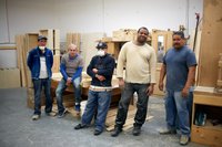 Five workers pose on the factory floor amidst partially finished pieces of wood furniture