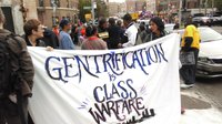 Protesters hold a white cloth banner with the words "Gentrification is Class Warfare"