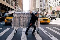 A man wearing a baseball hat guides a rack full of clothing through a crosswalk in front of taxi cabs paused at the red light.