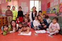 Teachers and children gather on the floor of a classroom with pink walls adorned with colorful artwork