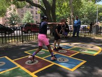 Children playing hopscotch under a shade tree while an older woman watches. Brick apartment buildings are visible through the trees in the background.