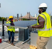 Two contractors wearing reflective vests and hard hats carry a solar panel into place on the roof of a NYCHA building.