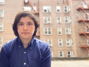 Photo of Lena Afridi wearing a blue button-down shirt, posing in front of a New York City apartment building