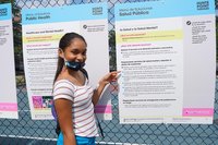 Girl wearing a face mask points to a poster titled "Public Health/Salud Publica" which is displayed with a series of posters on a chain-link fence