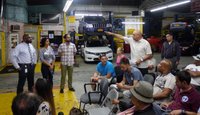 Three city agency representatives stand in front of small group seated in chairs in an auto repair garage. One man stands among the group speaks and points in the direction of the officials.