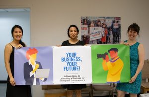 Three women hold a banner picturing a photographer and a business person holding a laptop with blue text reading "Your Business, Your Future"
