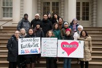 Group poses on the steps of City Hall holding signs that read: "Basements Keep East New York Safe-Stable-Affordable", and "Basement Apartments=Affordable, Safe, Stabilizes Homeownership, Prevents Homelessness, Protects Tenants, a place to call home"