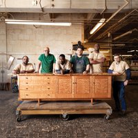 Six workers stand in a row behind a long horizontal wood cabinet inside a wood shop.