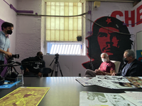 Film crew interviews two elderly men sitting under a large Che Guevara mural.