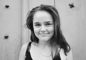 Black and white photo. Headshot; smiling young woman with brown hair and black tank top standing in front of wood-planked wall