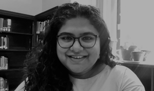 Black and white. Headshot. Young woman with dark, curly hair and glasses, white shirt, smiling. Background library setting.