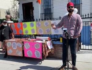 Man in plaid shirt with beard and sunglasses speaks into microphone with neon posters lining an iron fence behind him.