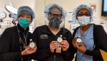 Three healthcare workers wearing scrubs and masks hold small bottles of hand sanitizer