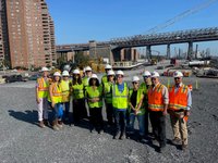 A group wearing reflective vests poses at a large construction site with heavy equipment and Williamsburg Bridge in the background